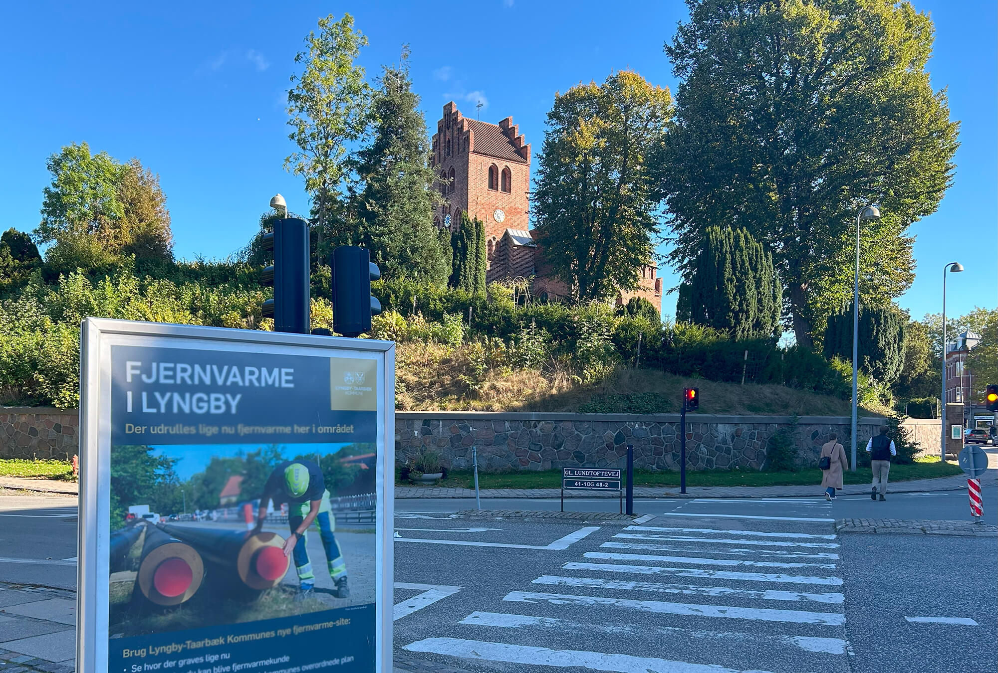 Byggeskilt foran vejkryds med Lyngby Kirke i baggrunden